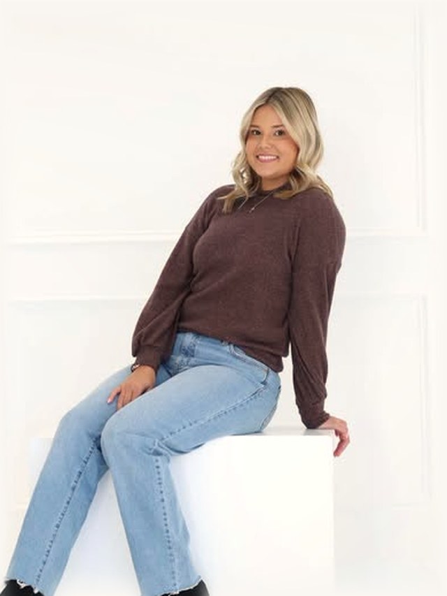 Caydence, hairstylist, seated in a brown knit and jeans against the salon&rsquo;s white studio wall.