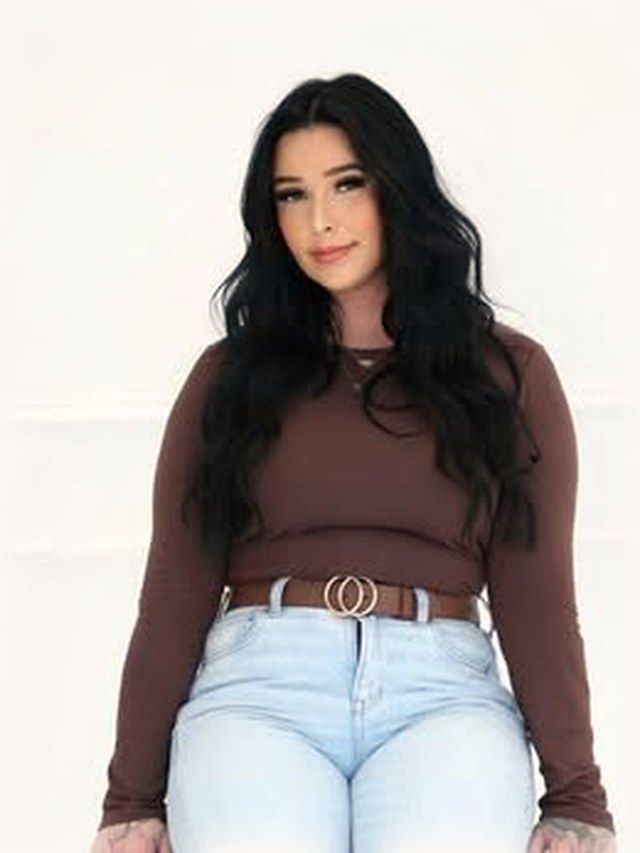 Sierra, hairstylist, in a brown top and jeans against the salon&rsquo;s white studio wall.